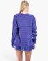 A woman viewed from the back wearing a textured, ribbed knit sweater in a medium purple shade with a fringed hem, set against a plain white background.