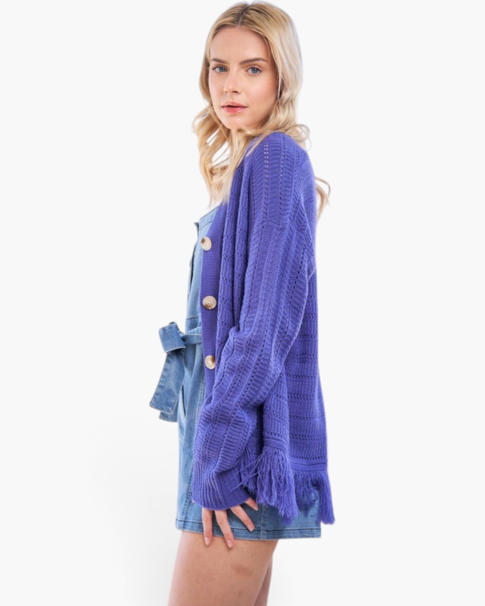A woman modeling a textured purple knit cardigan with button closures and fringe details, worn over a denim dress against a plain white background.