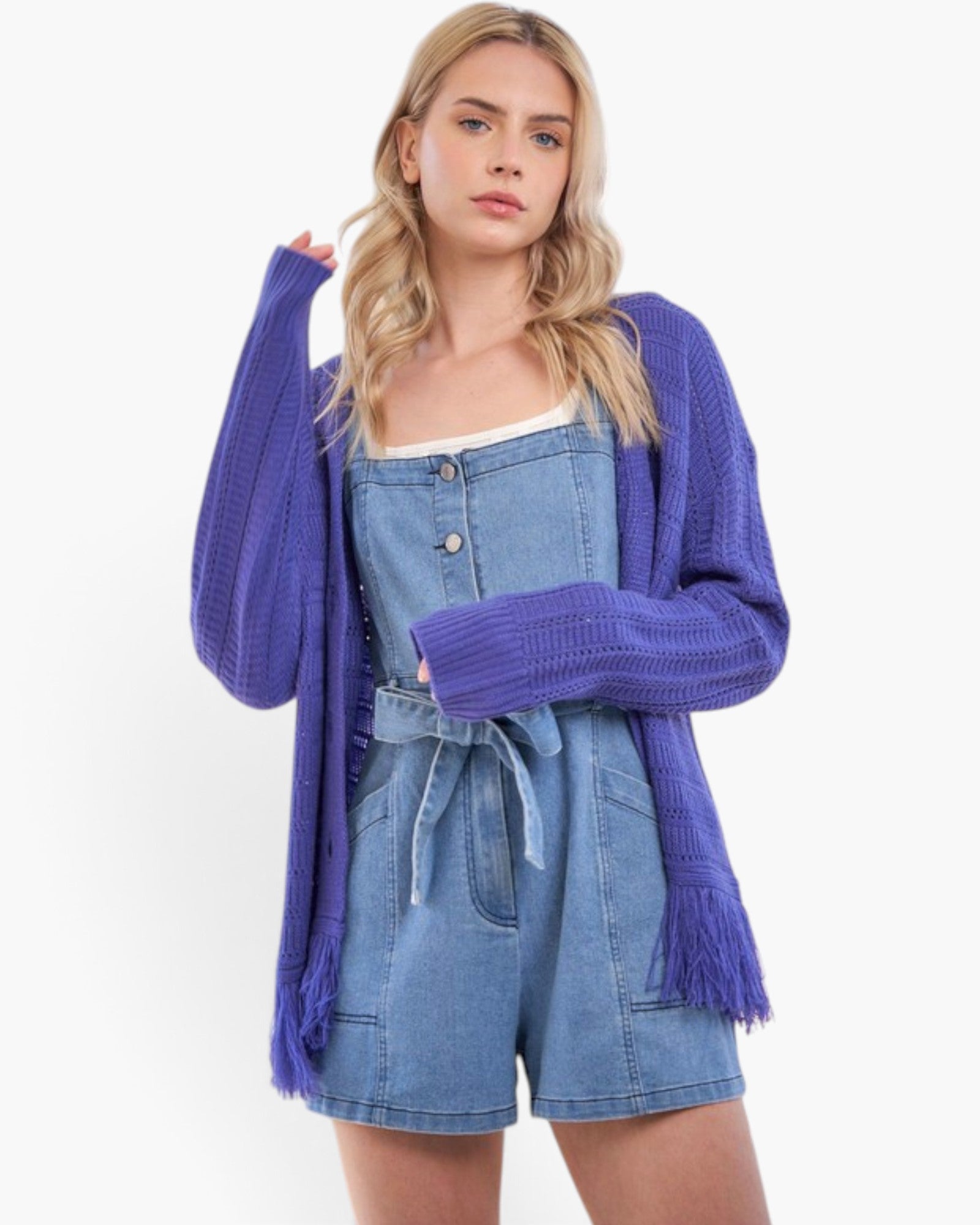 A woman modeling a vibrant purple knit cardigan with fringe details over a light blue denim romper, set against a plain white studio background.