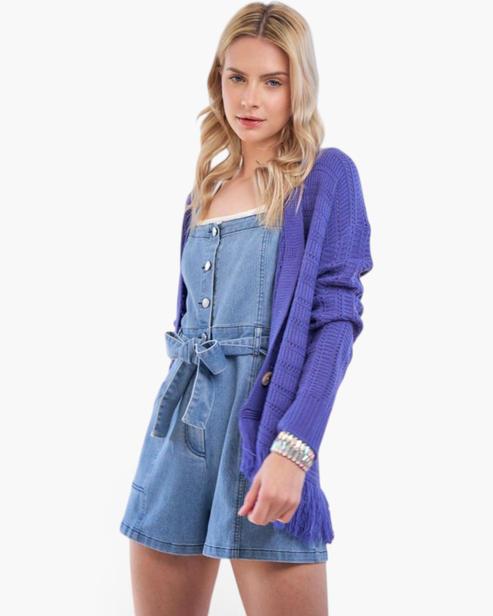 A blonde model wearing a purple knit cardigan with fringe over a blue denim mini dress, posed against a solid white studio background.