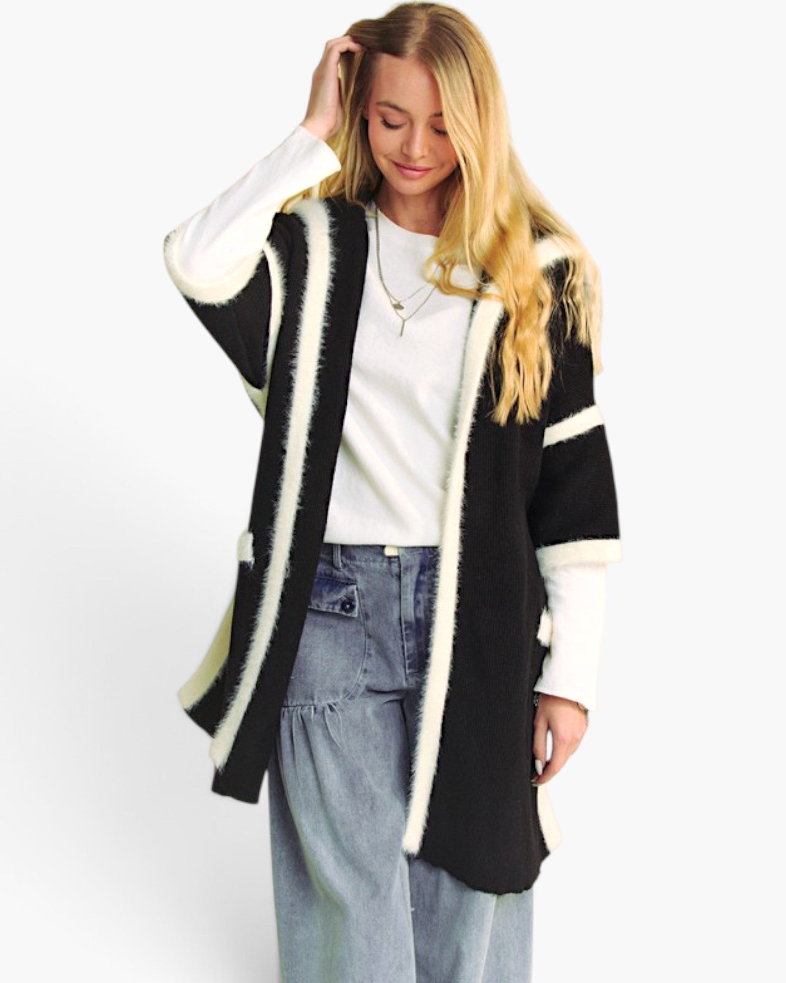 A woman modeling a black fuzzy open-front cardigan with cream-colored trim, worn over a white top and light-wash jeans against a plain white background.