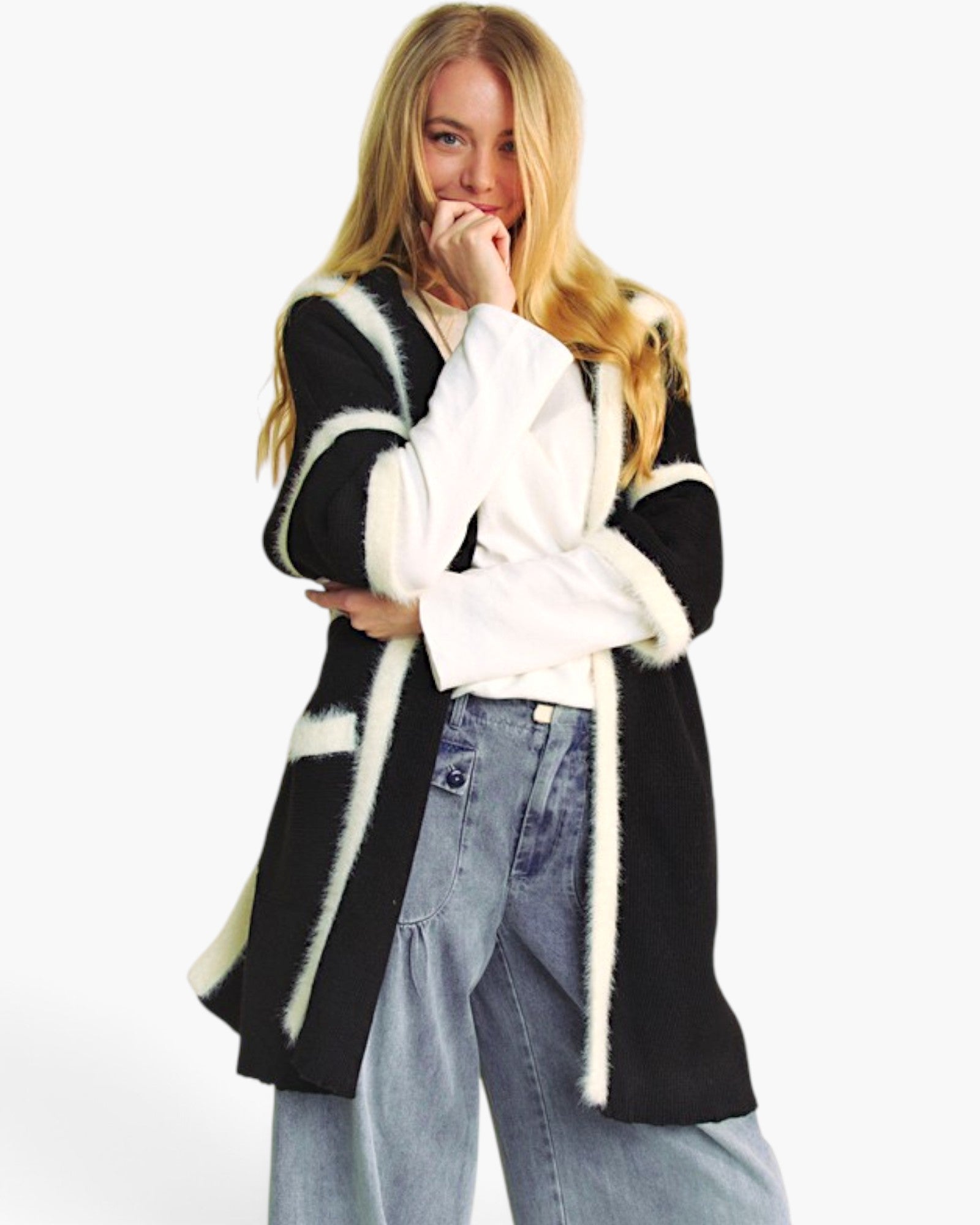 A blonde woman posing in a black fuzzy open-front cardigan with contrasting cream-colored trim, worn over a white long-sleeve top and light-wash blue jeans against a plain white studio background.