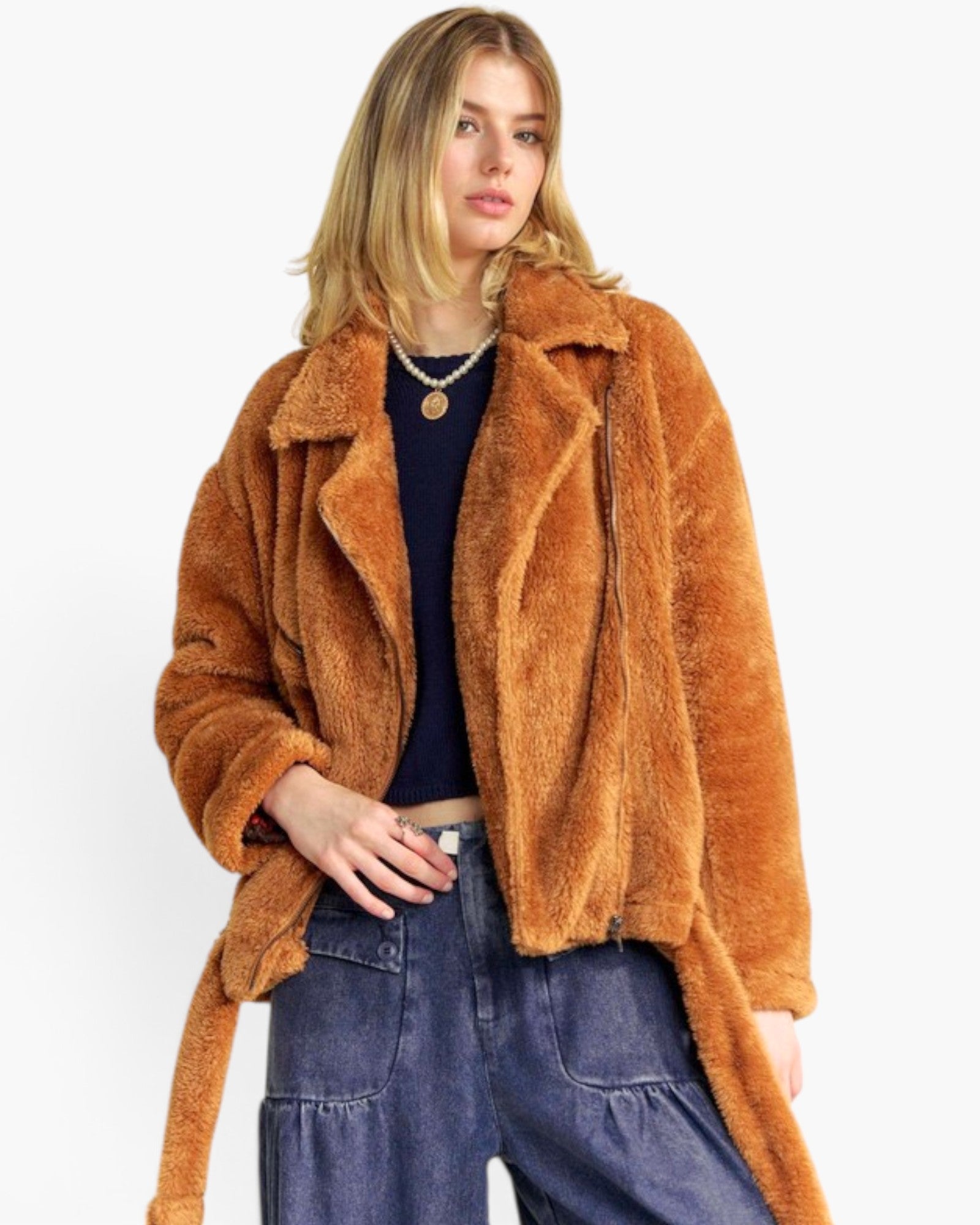 A woman modeling a brown plush teddy-fleece jacket with a collar and tie belt, worn over a dark top and blue jeans against a plain white background.