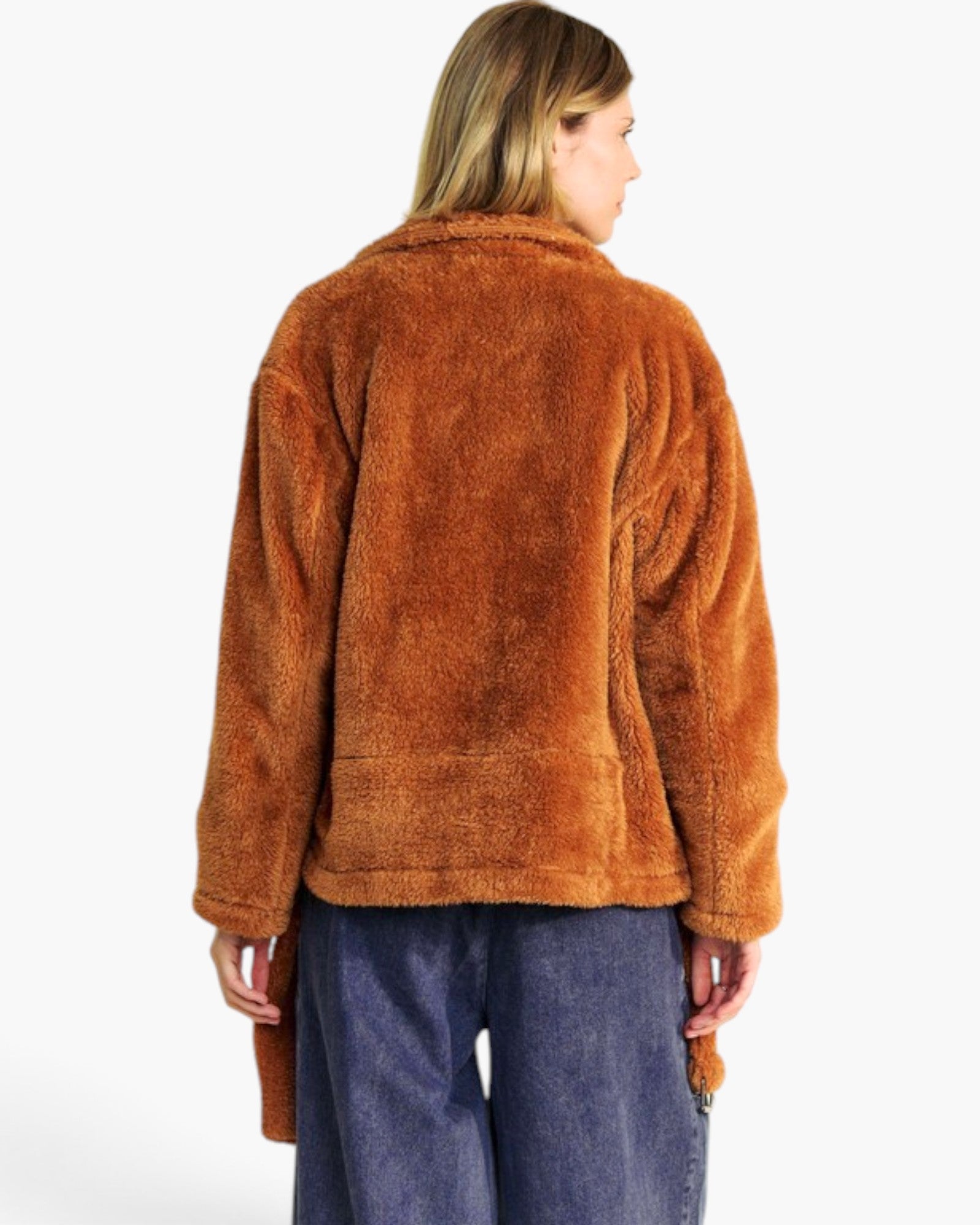 A woman shown from the back wearing a warm rust-brown sherpa fleece jacket and blue jeans against a plain white studio background.