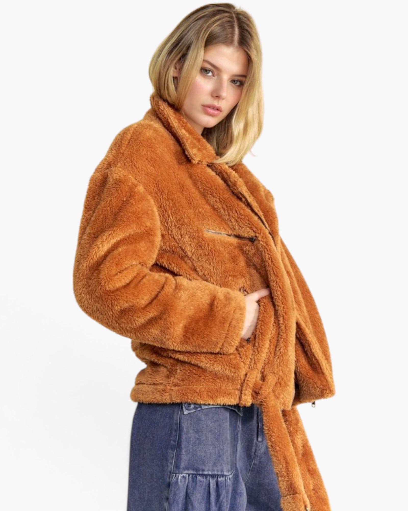 A woman wearing a cropped, warm brown sherpa fleece jacket with a collar and front zipper, paired with blue jeans, against a plain white background.