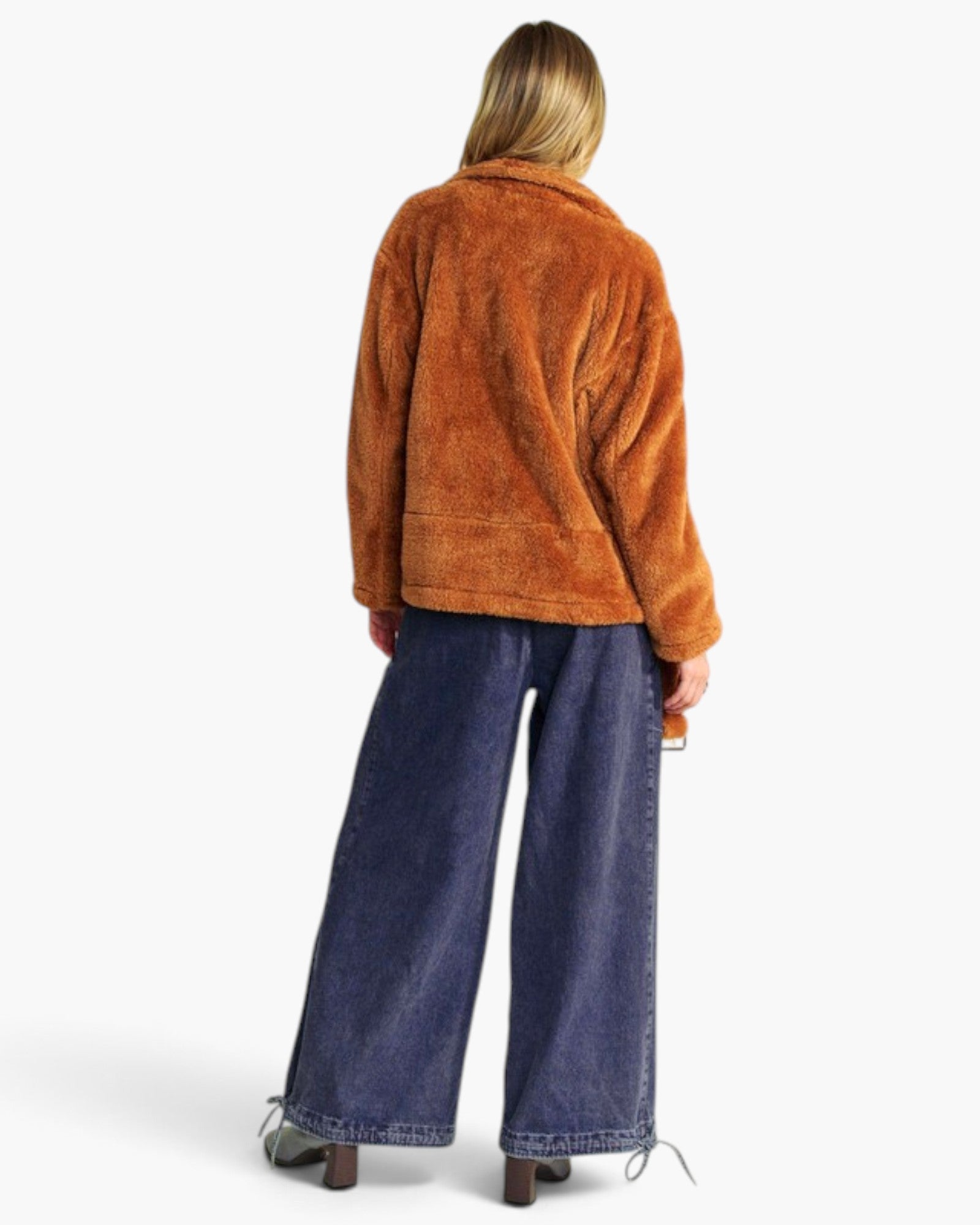 A woman shown from the back wearing a brown sherpa fleece jacket and wide-leg blue jeans, photographed against a clean white studio background.