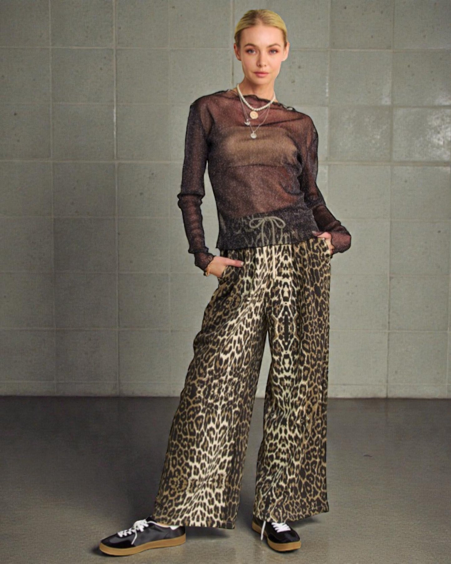 Woman wearing a sheer top and leopard print pants against a gray tiled wall.