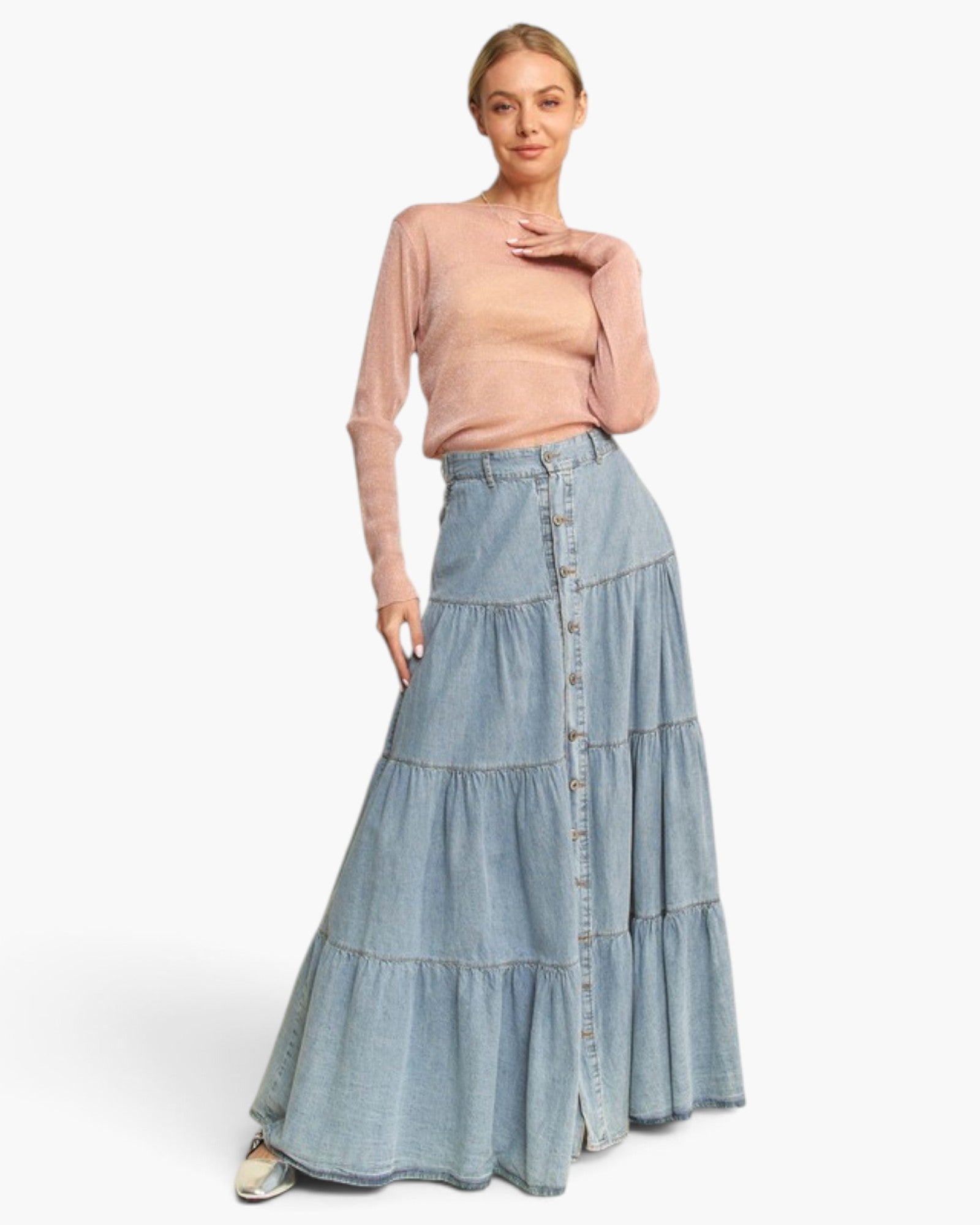 A woman modeling a light pink long-sleeve knit top and a high-waisted, tiered light blue denim skirt with a button-front closure, photographed against a clean white studio background.