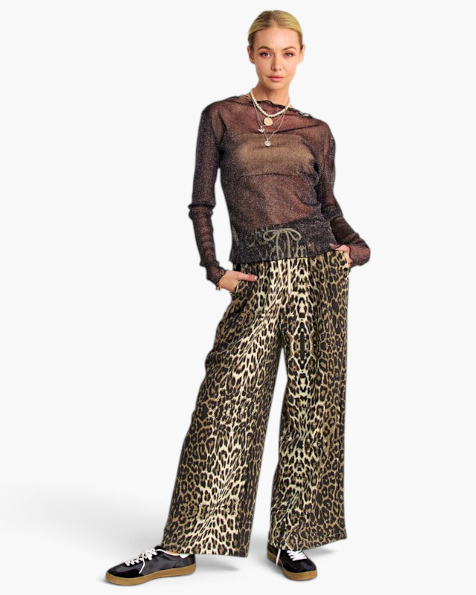 A model wearing a sheer brown long-sleeve top and wide-leg leopard print pants, paired with black sneakers, against a light gray studio background.