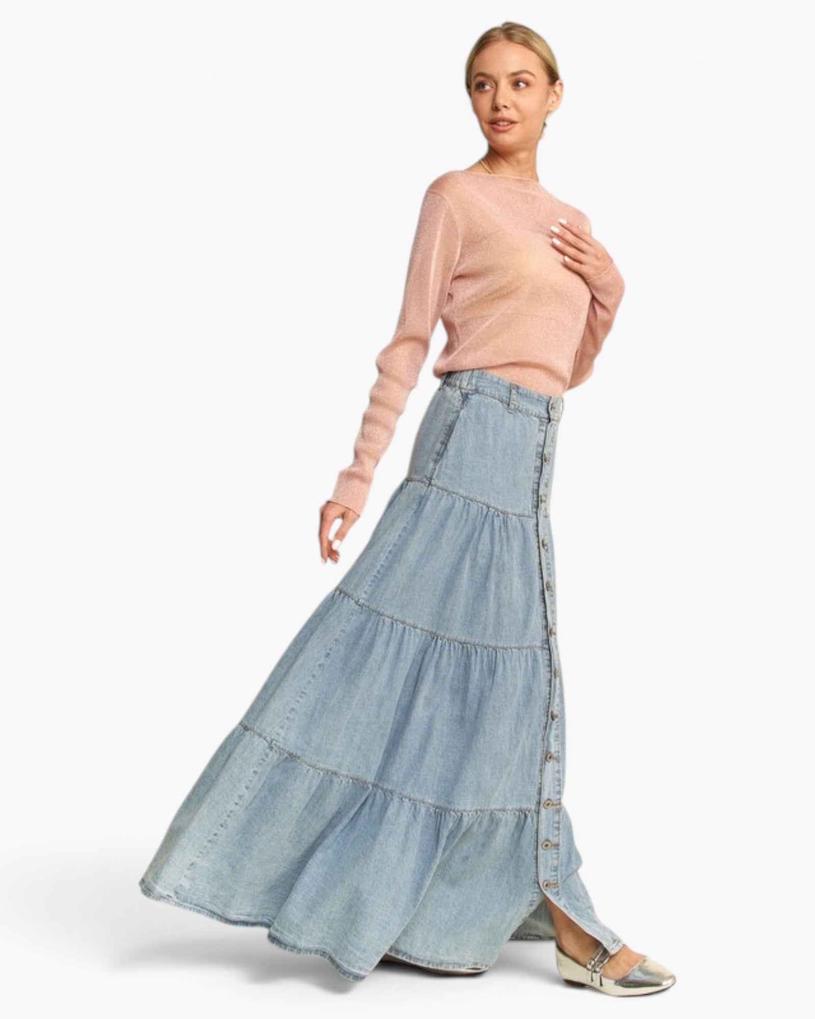 A woman modeling a light blue tiered denim maxi skirt with a buttoned side slit, paired with a sheer pale pink long-sleeve top, against a plain light gray studio background.