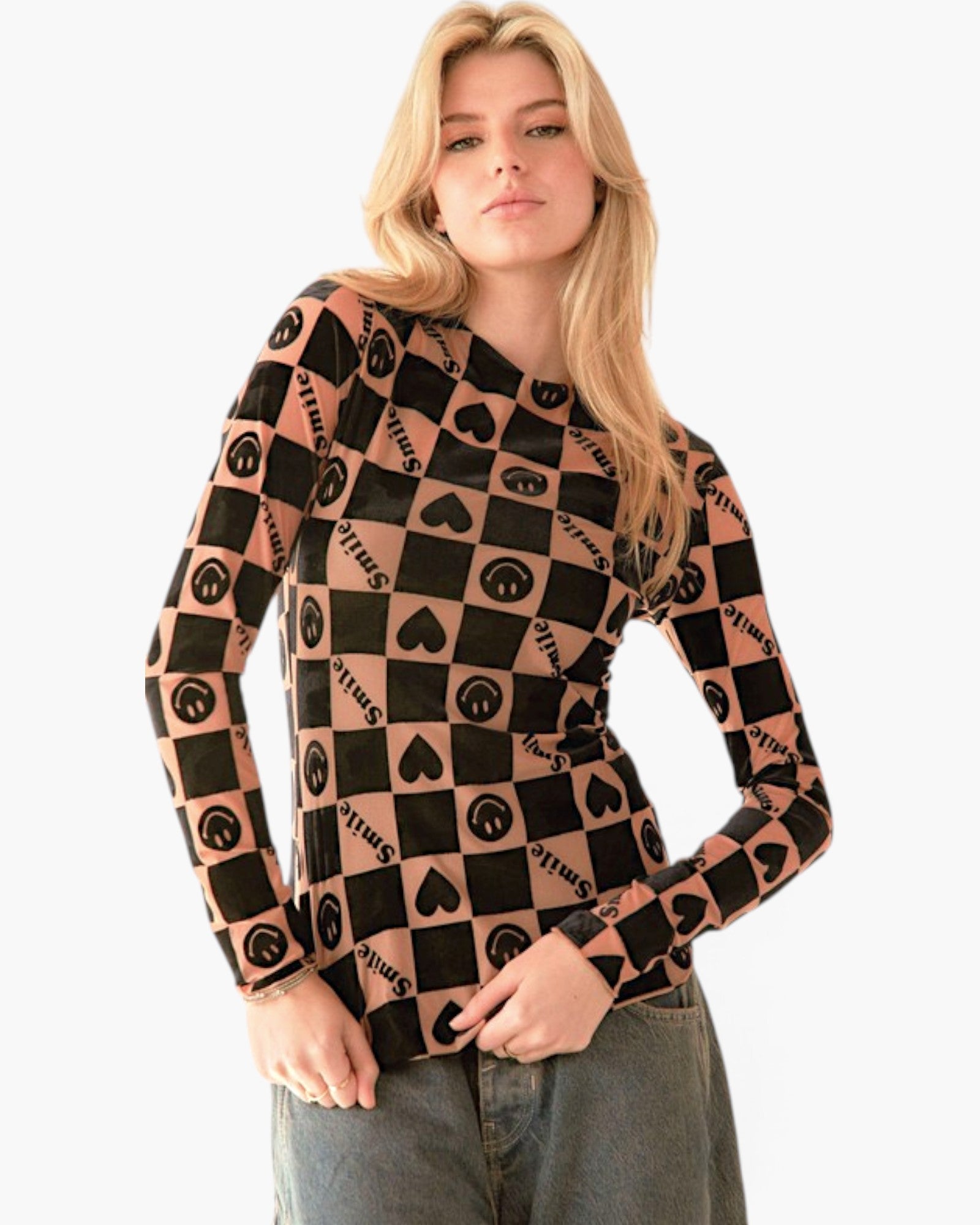 A woman modeling a long-sleeve knit top with a black and peach checkerboard pattern featuring peace signs and hearts, paired with gray jeans against a plain white background.