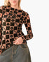 A woman wearing a long-sleeve sheer mesh top with a black and tan checkerboard pattern featuring hearts, smiley faces, and the word 'Smile' printed repeatedly, paired with a denim skirt against a plain white background.