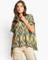 A woman modeling a short-sleeve knit top with a geometric Southwestern pattern in olive green, turquoise, mustard yellow, and beige, paired with khaki cargo pants against a light gray studio background.