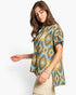 A woman wearing a short-sleeve knit top with a colorful geometric Southwestern pattern in teal, yellow, beige, and brown, paired with beige pants, against a plain white background.