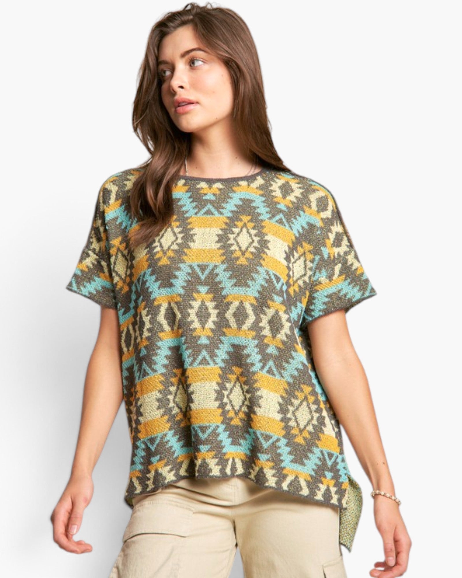 A woman wearing a short-sleeve knit top with a geometric Southwestern pattern in olive green, beige, turquoise, and mustard yellow, paired with khaki cargo pants, against a light gray studio background.