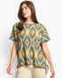 A woman wearing a short-sleeve knit top with a geometric Southwestern pattern in olive green, beige, turquoise, and mustard yellow, paired with khaki cargo pants, against a light gray studio background.