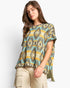 A woman modeling a short-sleeve knit poncho top with a colorful geometric Southwestern pattern in teal, yellow, beige, and gray, paired with khaki cargo pants against a plain white studio background.