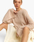 A woman wearing a chunky knit beige sweater with dramatic draped sleeves and a cream pleated skirt, posed against a clean white studio background.