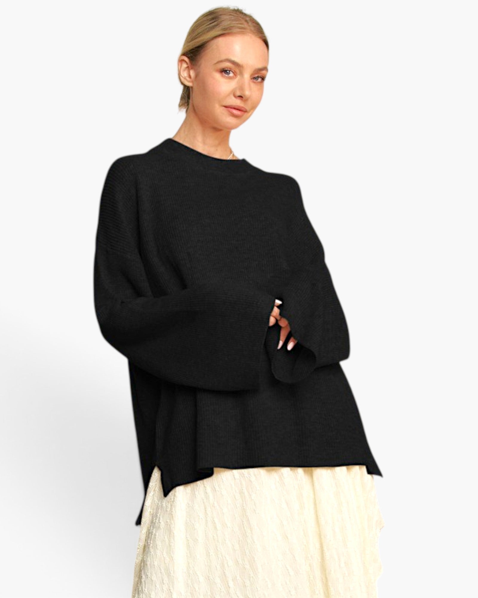 A woman modeling a black knit sweater with wide bell sleeves and a draped hem, paired with a cream textured skirt, against a plain light gray studio background.