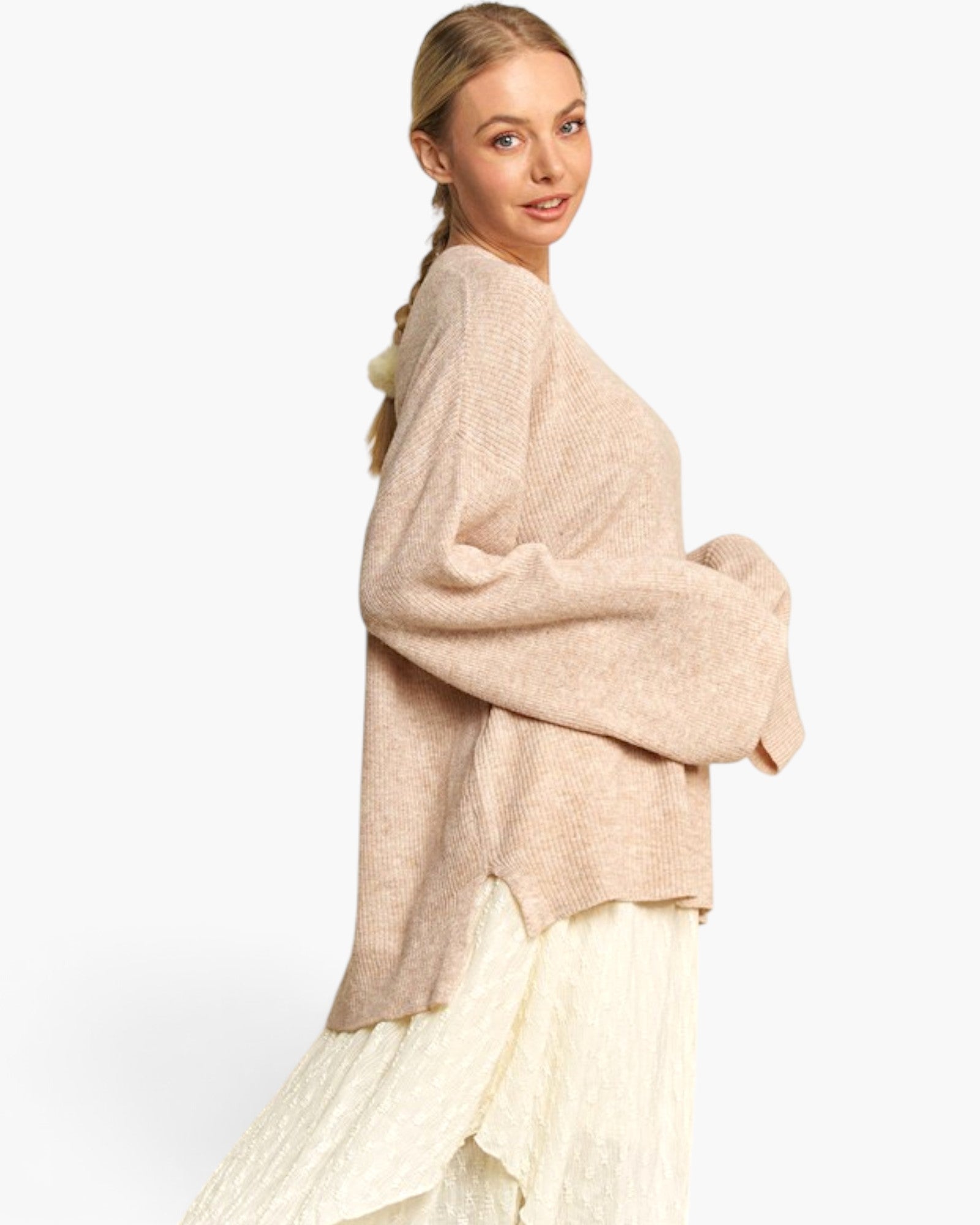 A woman wearing a loose, oversized knit cardigan in a light beige or blush tone, paired with a cream pleated skirt, photographed against a clean white studio background.