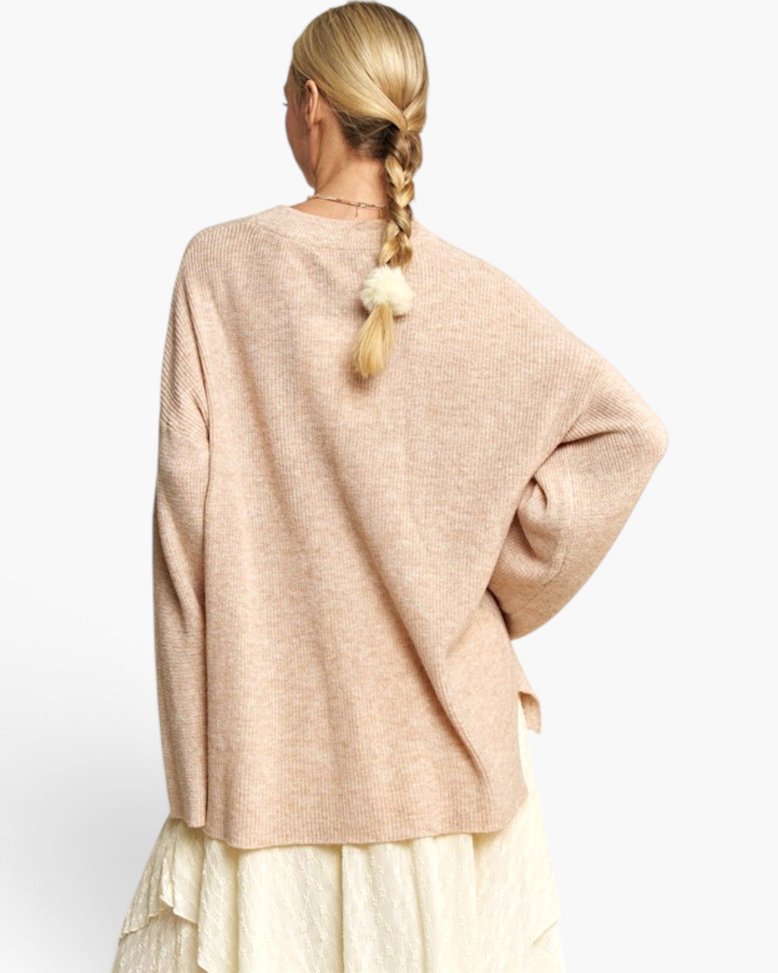 A woman shown from the back wearing a loose, oversized knit sweater in a light warm beige or blush tone, paired with a cream skirt. The sweater has a crew neck and wide sleeves, with a textured knit pattern.
