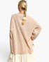 A woman shown from the back wearing a loose, oversized knit sweater in a light warm beige or blush tone, paired with a cream skirt. The sweater has a crew neck and wide sleeves, with a textured knit pattern.