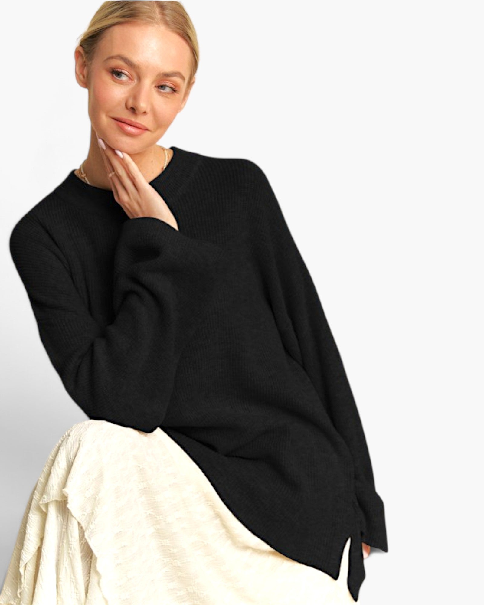 A woman wearing an oversized black knit sweater and a cream-colored skirt, posing against a solid light gray background.