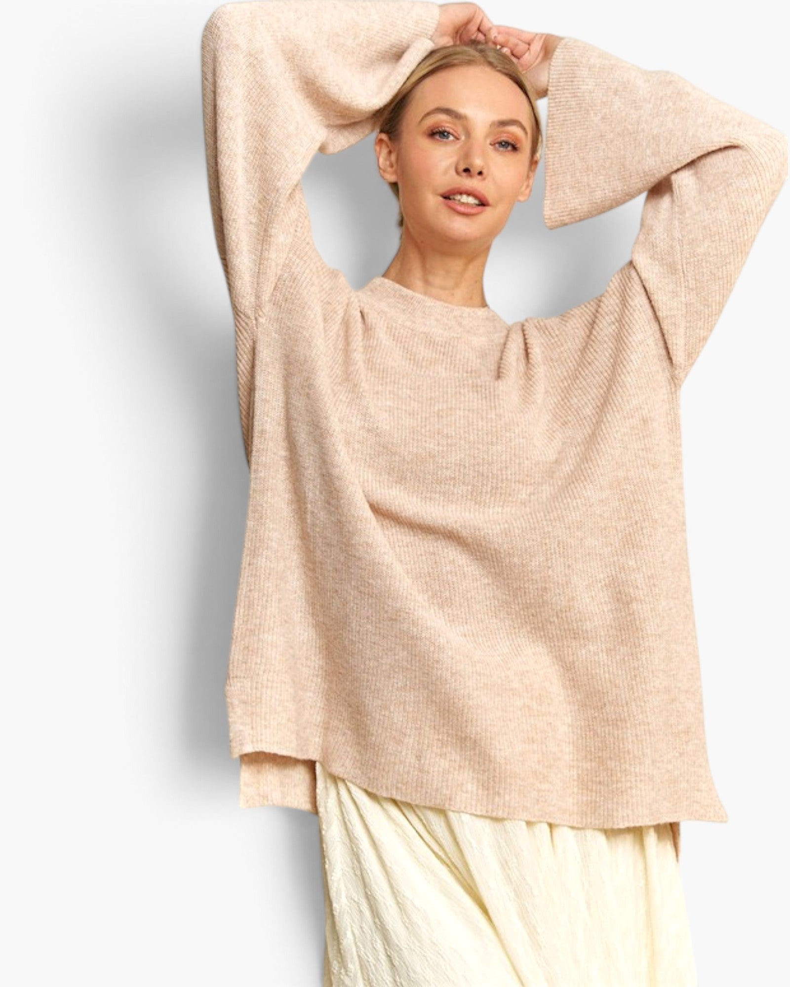A woman wearing a loose, oversized knit sweater in a light beige or cream color, paired with light cream pants, posing with her arms raised against a clean white background.