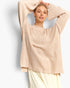 A woman wearing a loose, oversized knit sweater in a light beige or cream color, paired with light cream pants, posing with her arms raised against a clean white background.