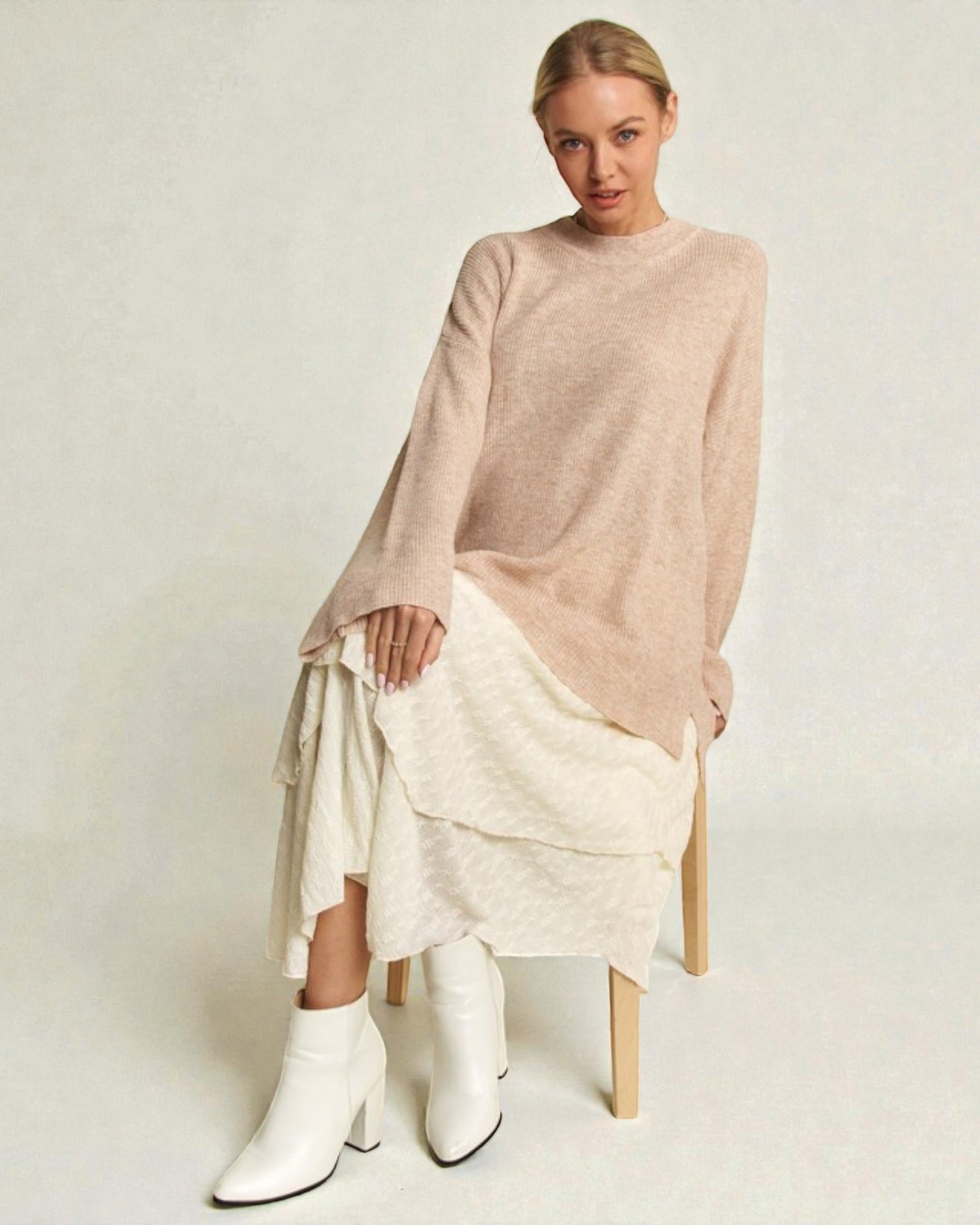 Woman wearing a beige sweater and white skirt with white boots on a light background