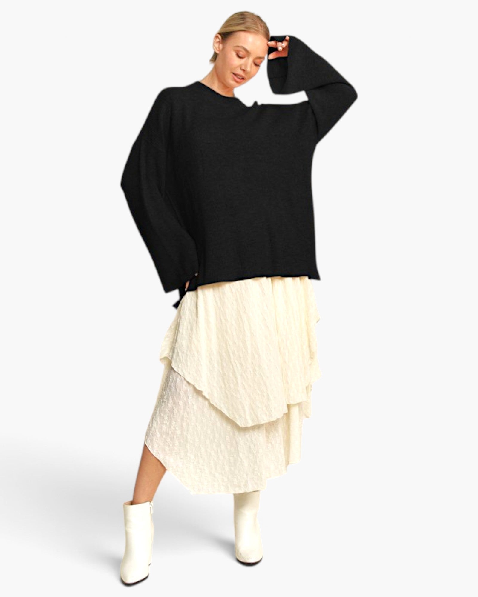A woman modeling a black oversized knit sweater with wide sleeves, paired with a cream ruffled midi skirt and white ankle boots, against a light gray studio background.