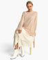 A woman seated on a wooden chair wearing a loose beige knit sweater, a cream textured skirt, and white ankle boots against a clean white studio background.