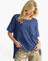 A woman wearing a textured blue short-sleeve top with a front pocket and metallic gold pants, photographed against a clean white background.