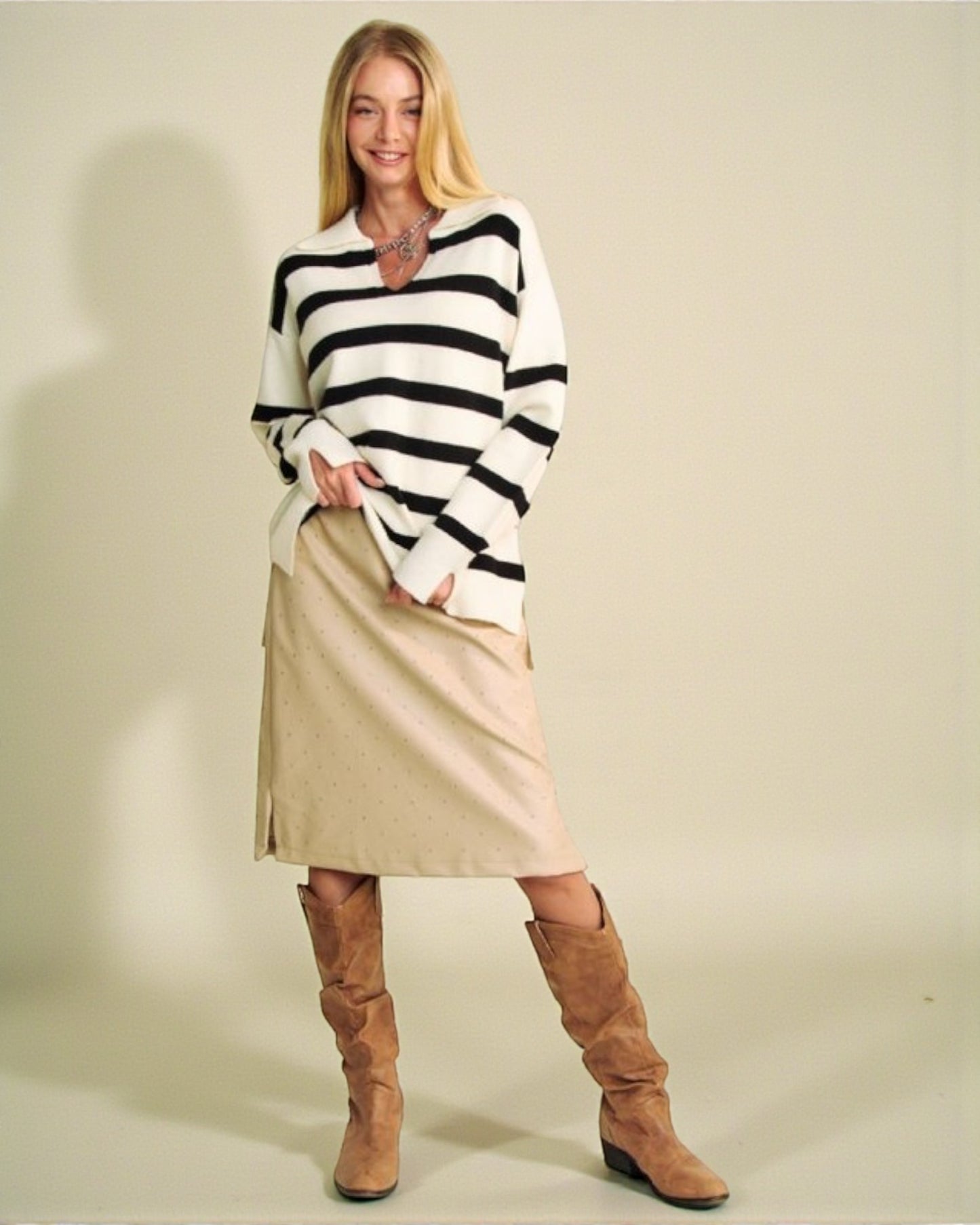 Woman wearing a black and white striped sweater, beige skirt, and brown knee-high boots against a plain background.