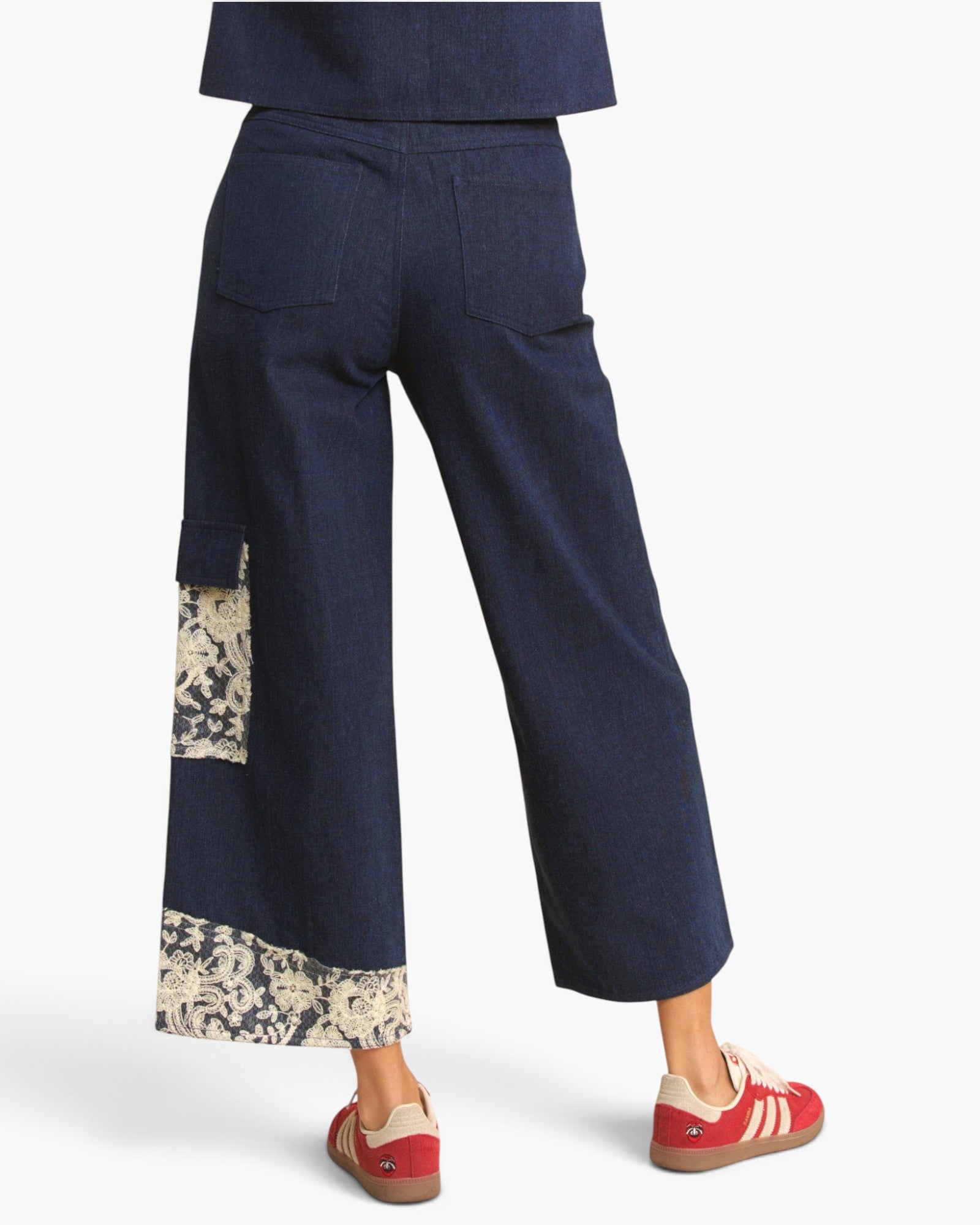 A person wearing dark navy wide-leg cropped pants with beige floral patchwork details on the lower legs, paired with red sneakers, shown from the back against a light gray studio background.
