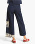 A person wearing dark navy wide-leg cropped pants with beige floral patchwork details on the lower legs, paired with red sneakers, shown from the back against a light gray studio background.