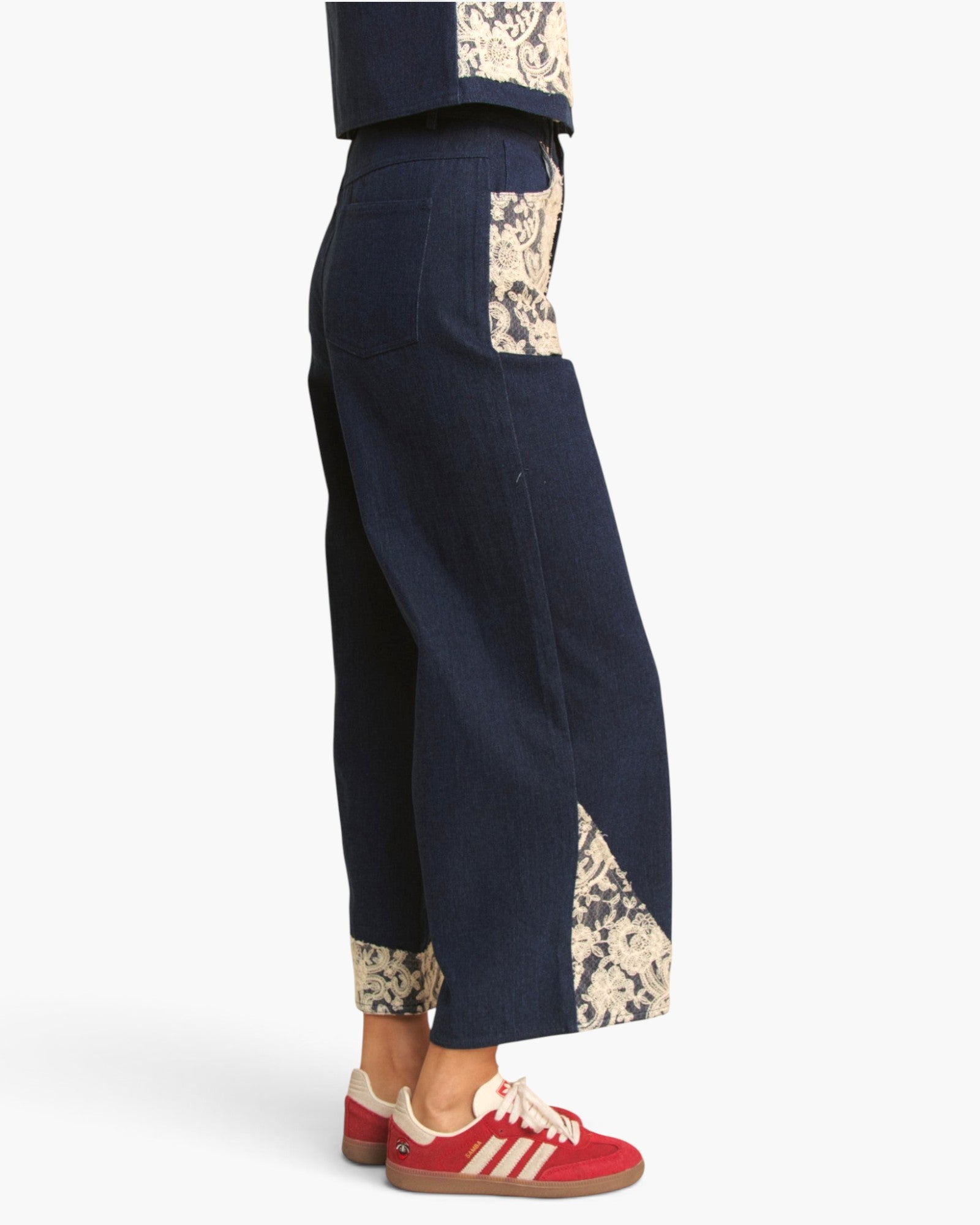 A person shown from the waist down wearing dark navy wide-leg cropped pants with beige floral patterned panels at the pockets, hem, and cuffs, paired with red sneakers on a light gray background.