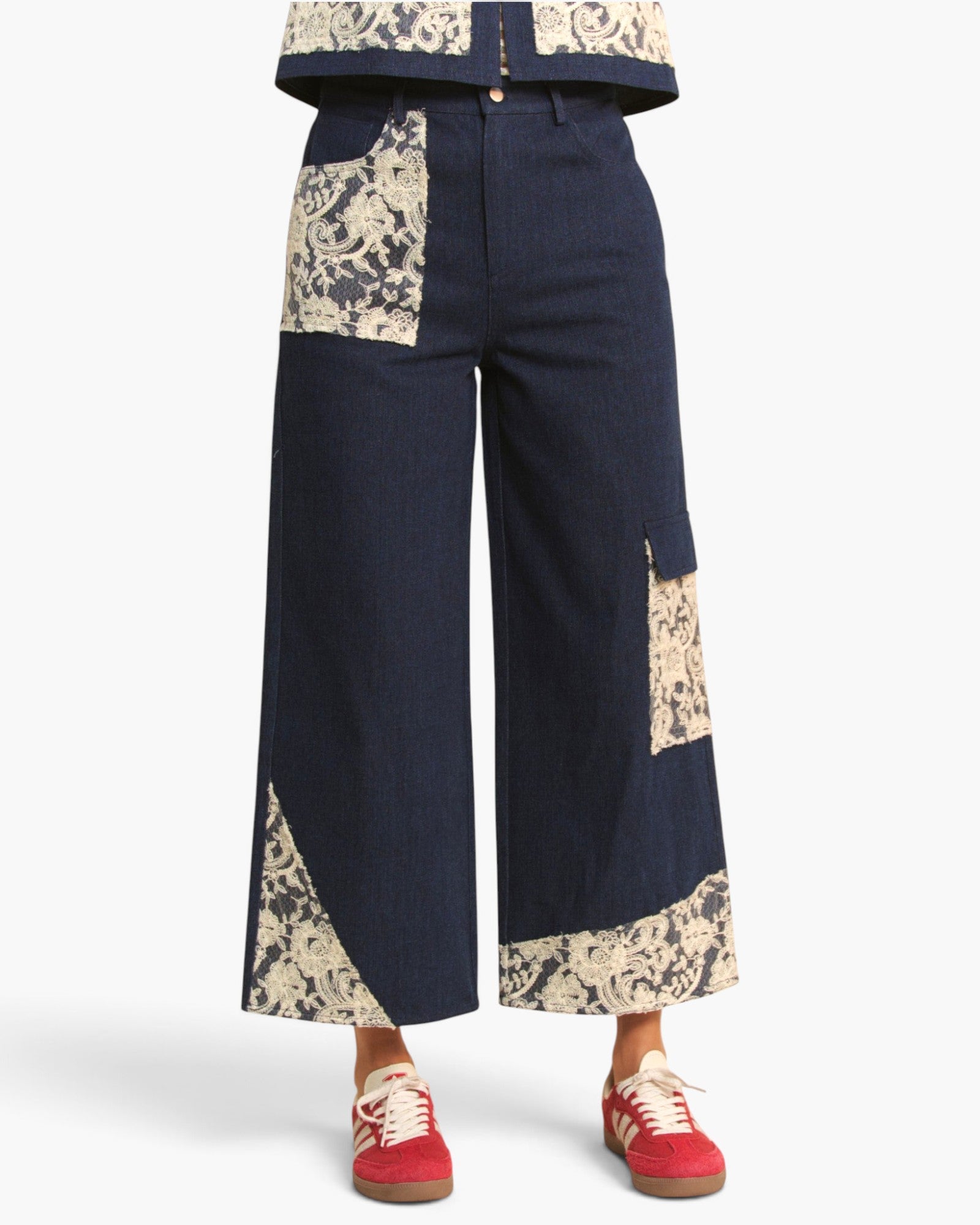 A person wearing dark navy wide-leg patchwork pants with beige floral print inserts, paired with red sneakers, shown from the waist down on a light gray studio background.