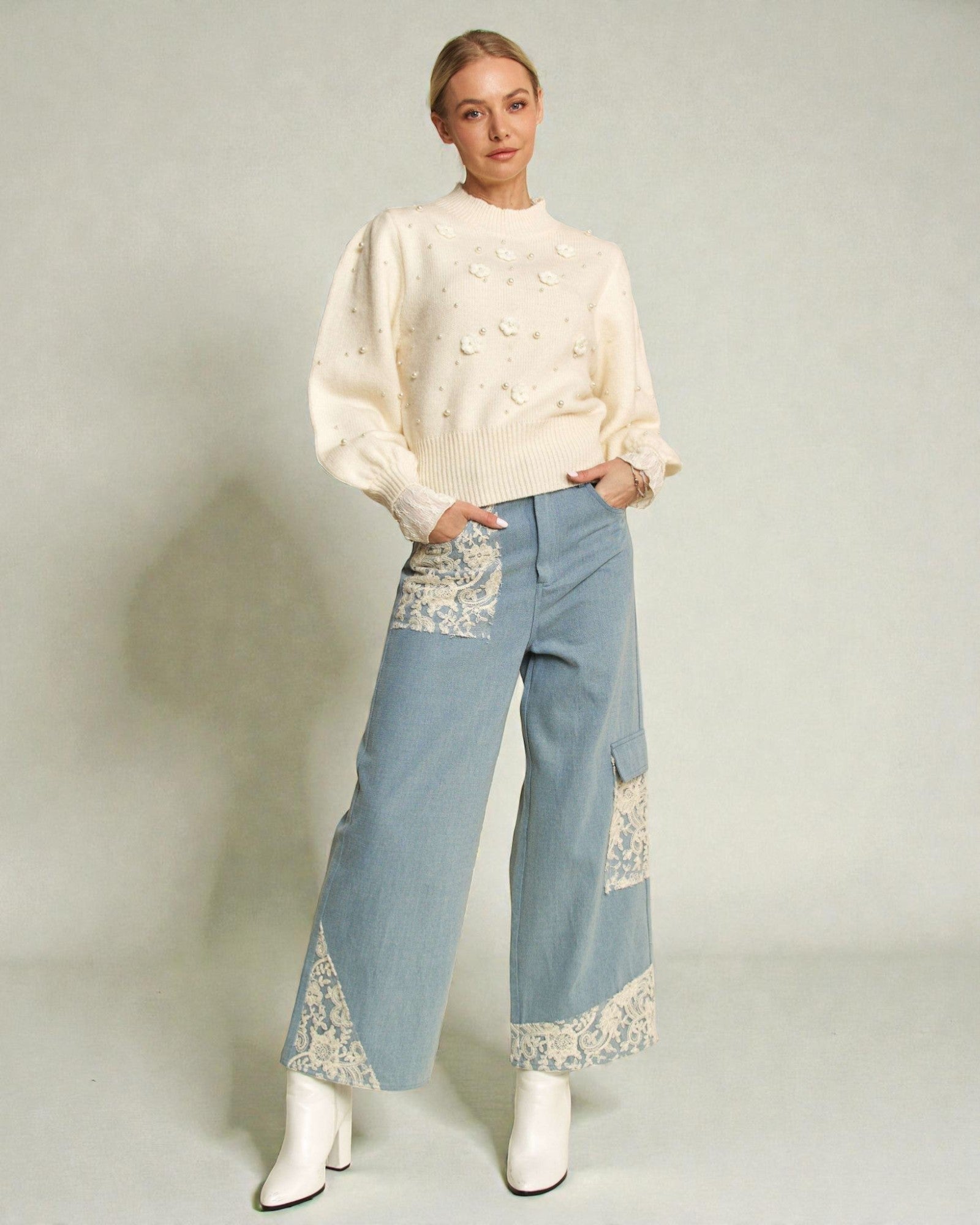 Woman wearing a cream sweater and light blue jeans with lace details on a plain background
