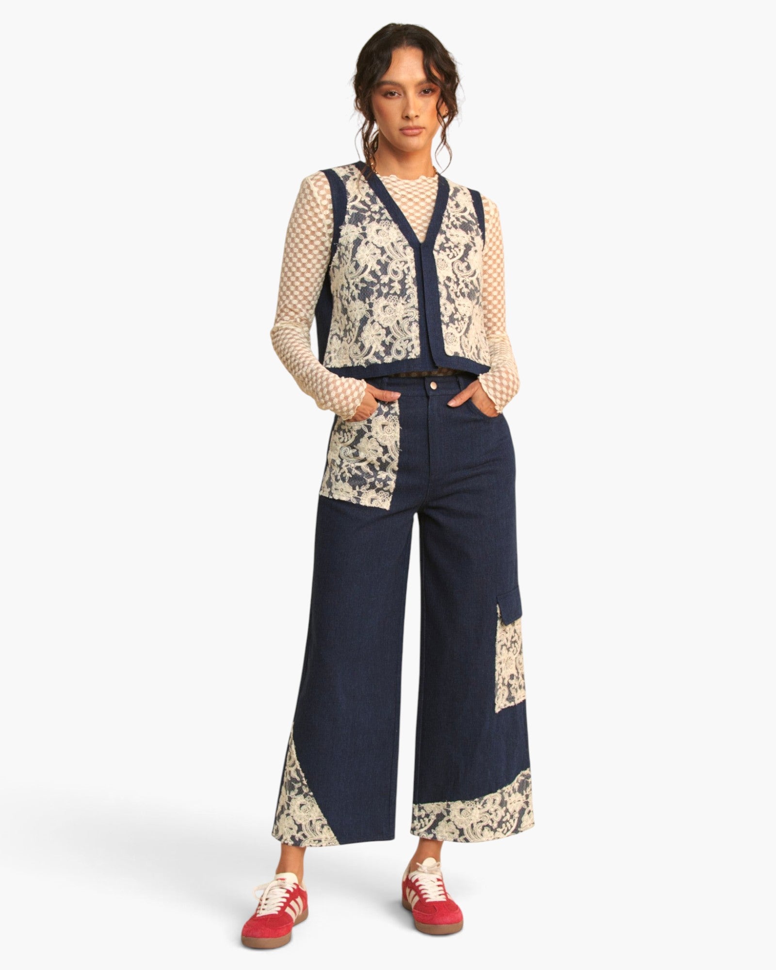 A model stands against a light gray studio background wearing a patchwork knit vest and wide-leg pants set in navy and cream, paired with red sneakers.