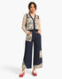 A model stands against a light gray studio background wearing a patchwork knit vest and wide-leg pants set in navy and cream, paired with red sneakers.