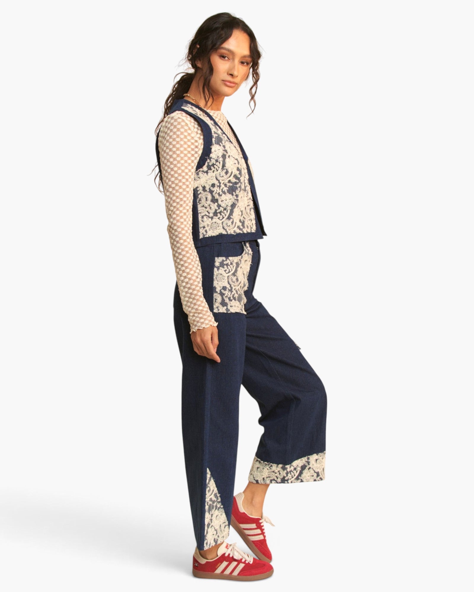 A model wearing a navy blue and beige patterned jumpsuit with a beige mesh long-sleeve shirt underneath and red sneakers, posed against a light gray studio background.