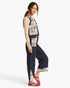 A model wearing a navy blue and beige patterned jumpsuit with a beige mesh long-sleeve shirt underneath and red sneakers, posed against a light gray studio background.