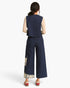 A model viewed from the back wearing a navy blue vest and matching wide-leg pants with beige patterned sleeves and hem details, paired with red shoes, against a plain white background.