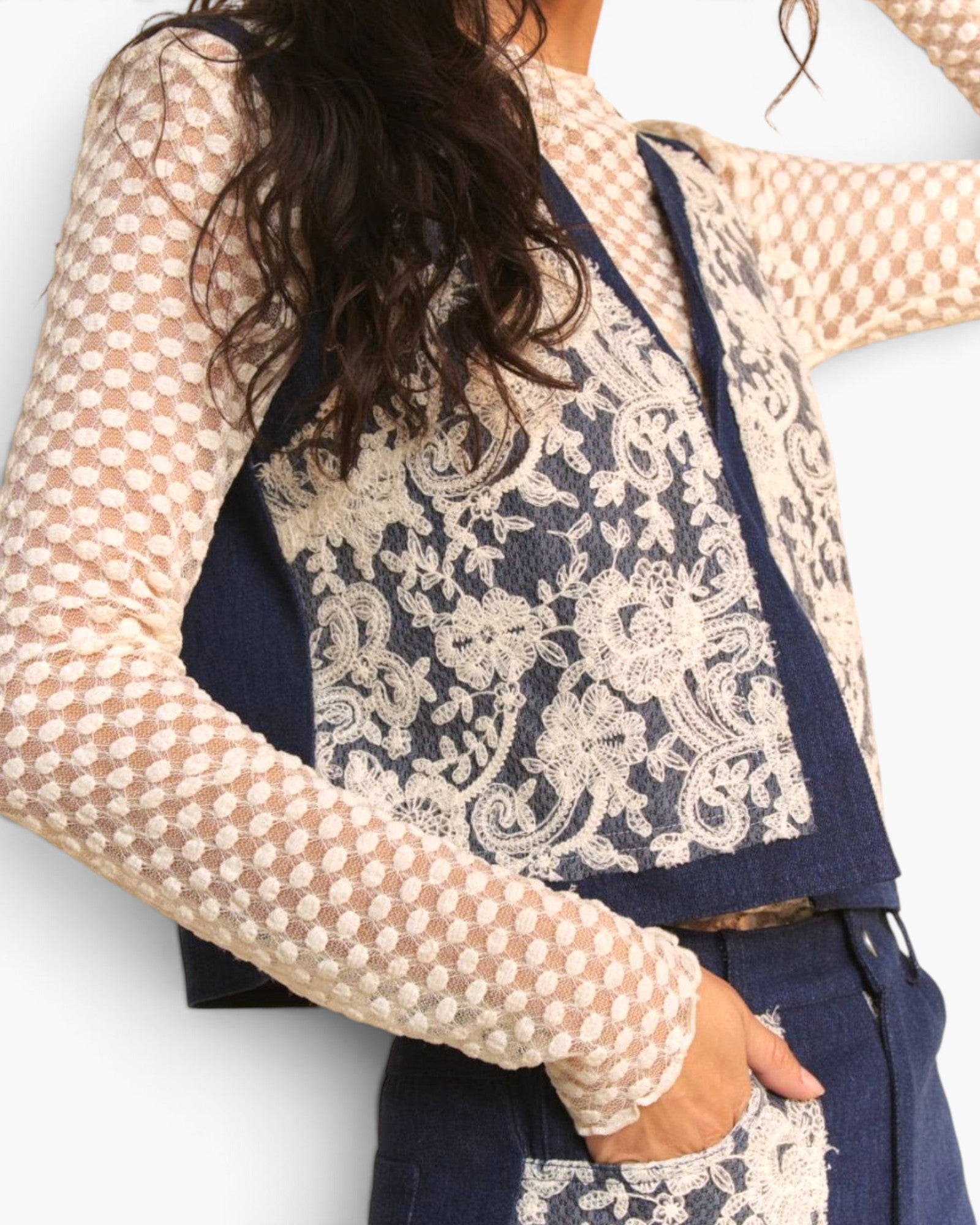 A close-up of a person wearing a navy blue denim vest with a large cream lace overlay panel, paired with a beige patterned long-sleeve top and dark blue jeans.