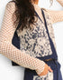 A close-up of a person wearing a navy blue denim vest with a large cream lace overlay panel, paired with a beige patterned long-sleeve top and dark blue jeans.