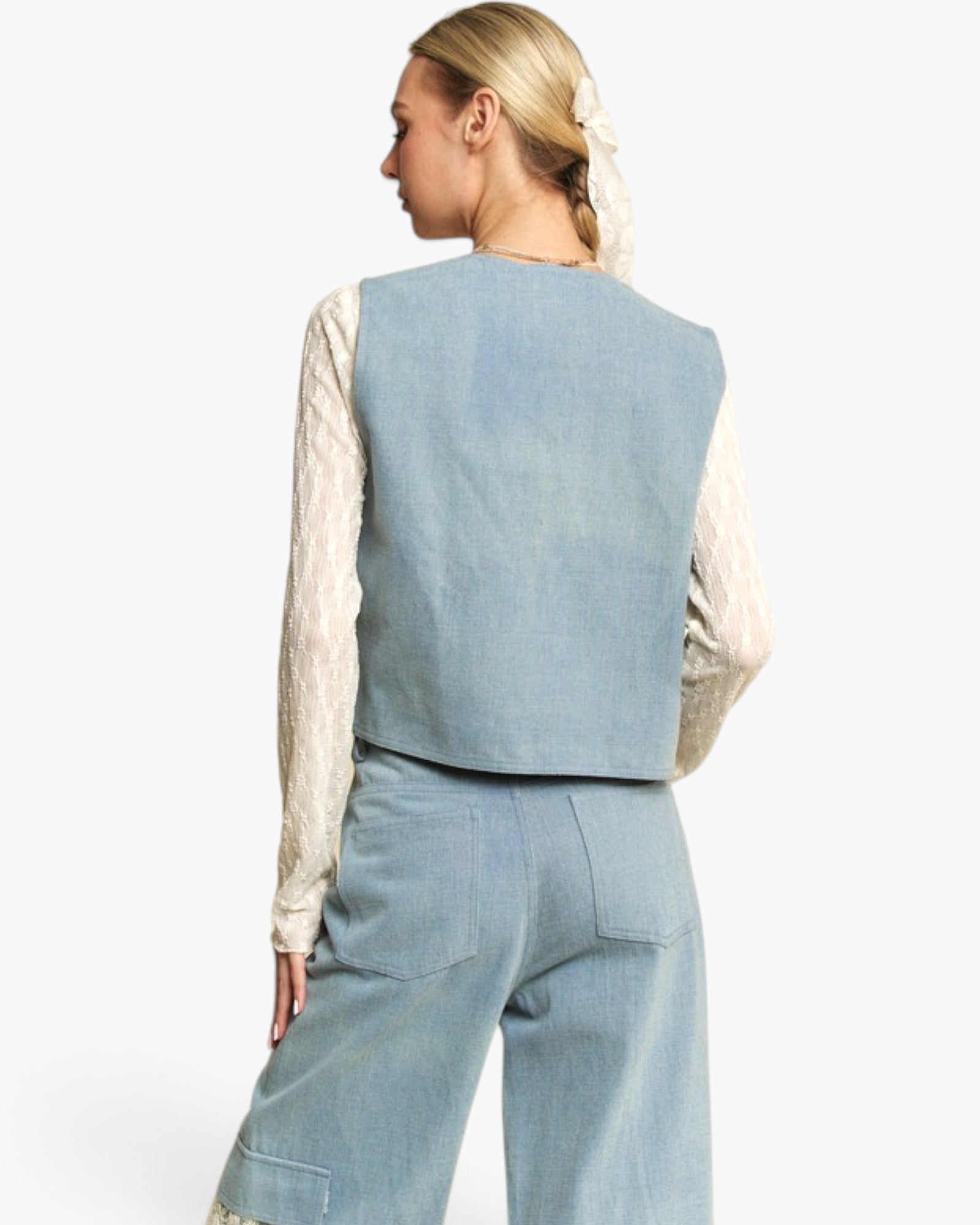 A woman shown from the back wearing a light blue linen vest over a cream lace long-sleeve top and matching light blue wide-leg trousers, set against a plain white studio background.