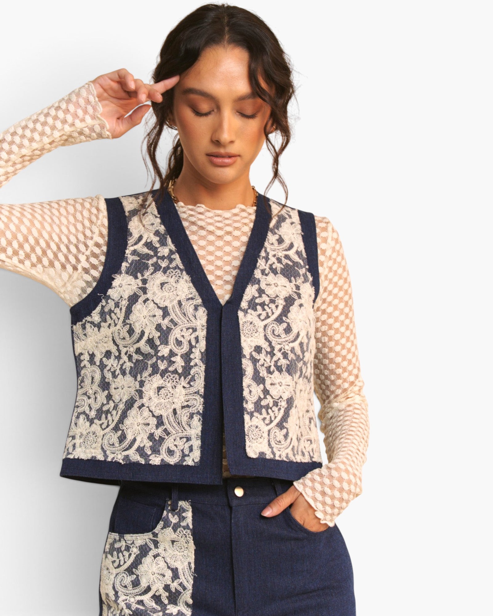 A woman wearing a navy blue and cream lace-patterned vest over a beige-and-white checkered long-sleeve top, paired with navy pants, against a light gray studio background.
