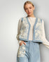 Woman wearing a floral-patterned cardigan over a white blouse with blue pants on a plain background