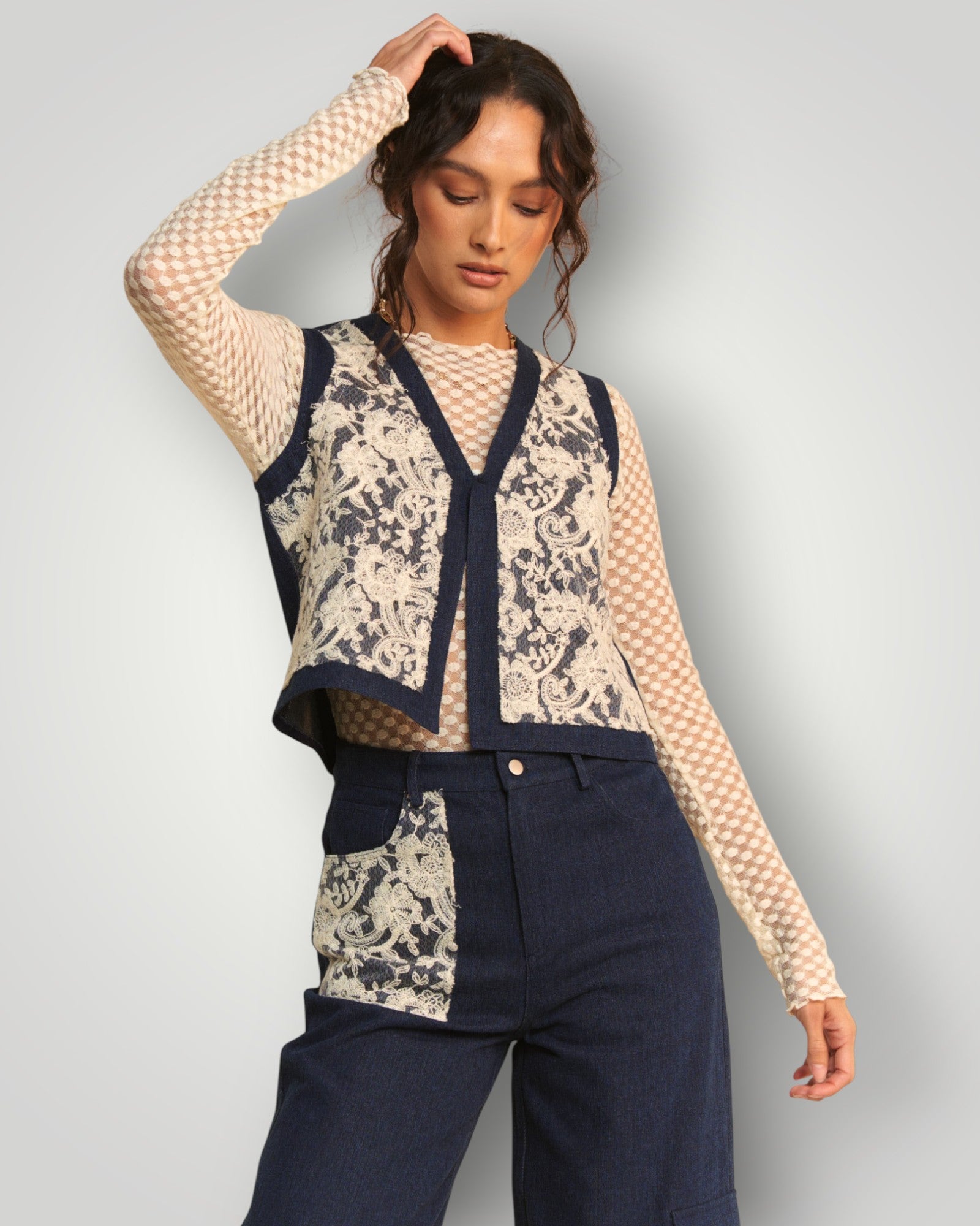 Woman wearing a patterned vest over a sheer top and dark pants on a plain background